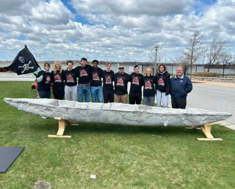 A group of people in matching shirts stands behind a concrete canoe, holding a pirate flag on grass—members of the American Society of Civil Engineers showcasing their team spirit outdoors.